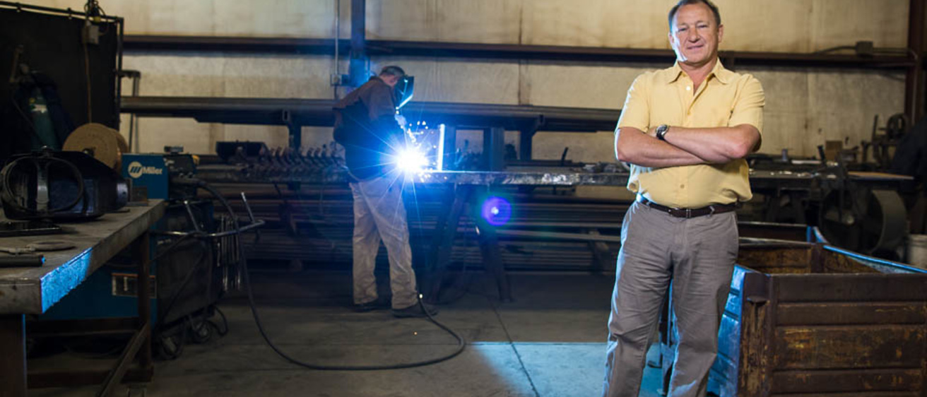 A man posing for a photo in a metal fabrication shop. Another man stands in the background welding a piece of metal with an arc welder.