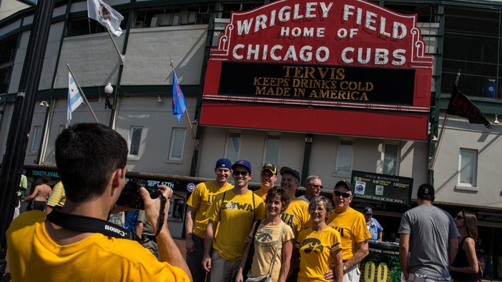 A man in a yellow t-shirt in the bottom left corner takes a photo of a group of people wearing Iowa Hawkeye and Cubs apparel in front of the Wrigley Field marquee.