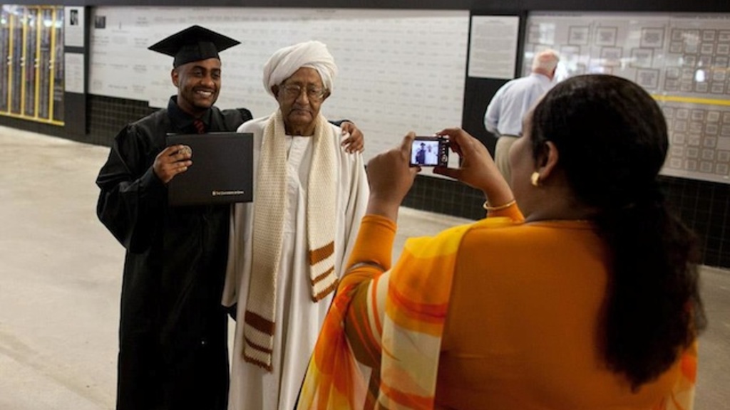 A woman takes a photo of a man in graduation garb alongside an older man.
